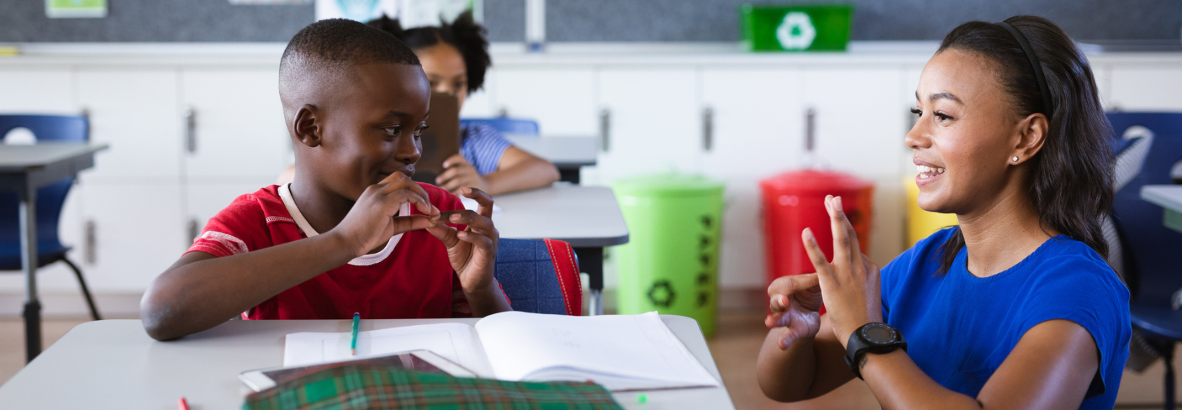 A teacher and a young student sit at a classroom table smiling and using sign language together. An open notebook and pencil are on the desk, and other students and colorful recycling bins are visible in the background.