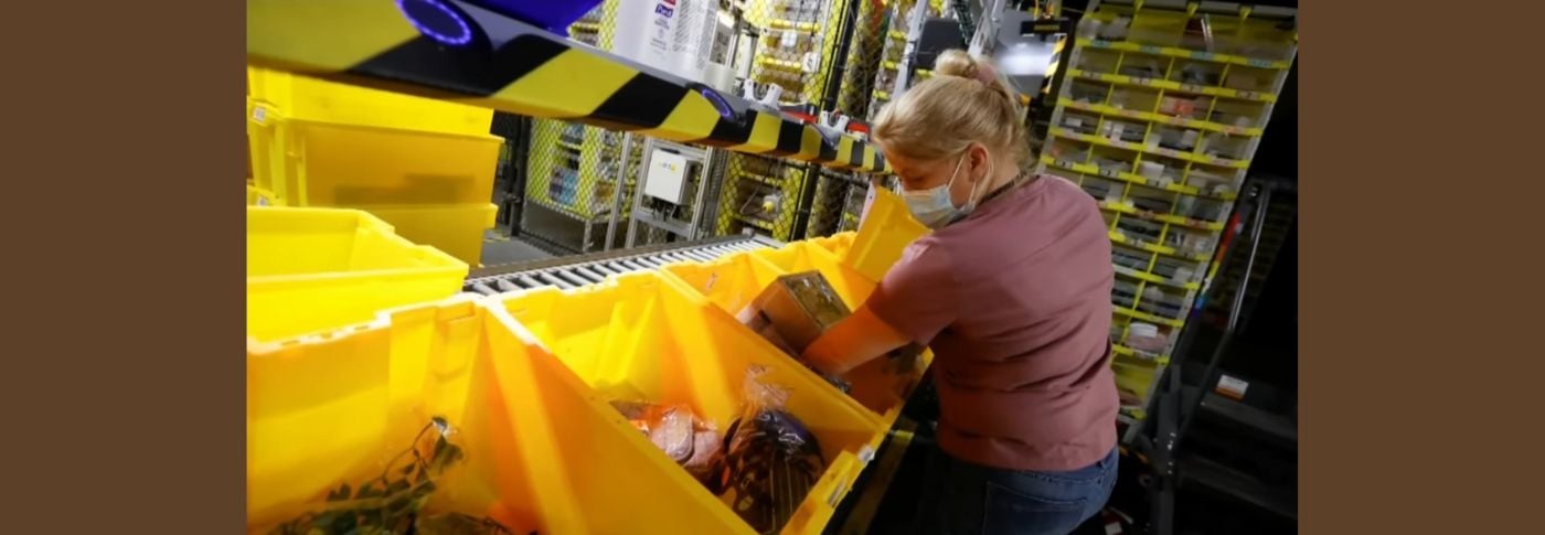 A warehouse worker wearing a face mask sorts packaged goods into yellow bins on a conveyor line. Despite full-time employment, many workers like her struggle to afford stable housing, highlighting the gap between wages and cost of living in the U.S.